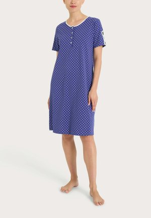 Donna che indossa un camicione da notte blu al ginocchio con pois bianchi, maniche corte e scollatura con bottoni, in piedi a piedi nudi.