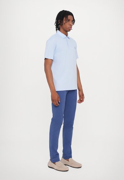 Selle helesinised lühikeste varrukatega polo särk, millel on tekstuuriga muster, koos tumedates sinistes slim-fit pükstega ja beezide mokassiinidega, seistes ühetoonilise tausta ees.