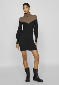 Abito in maglia nera con collo a drappeggio marrone e motivo a zigzag. Maniche lunghe a sbuffo e silhouette aderente. Indossato con stivali neri al ginocchio.