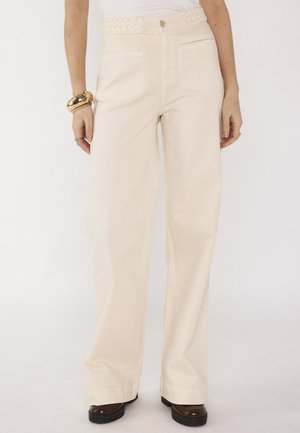 Donna che indossa pantaloni a gamba larga color crema a vita alta con cinturino intrecciato, top bianco, bracciale dorato e scarpe platform marroni su sfondo bianco.