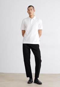 Hvit polo-skjorte med korte ermer og krage, kombinert med svarte bukser. Poloskjorten har en liten logo på brystet.