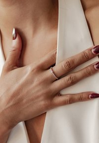 Bague en argent avec un petit diamant, présentant un anneau texturé. Une main avec des ongles rouge foncé repose sur un tissu crème.