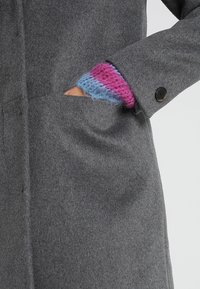 Šedý vlněný kabát s viditelnou boční kapsou a zapínáním na knoflíky. Částečně je vidět barevný pletený manžet v růžové a modré barvě.