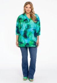 Losse tuniek in groen en blauwe abstracte print met een V-hals, driekwart mouwen, gecombineerd met flaired denim jeans en groene sandalen.