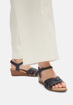 Granatowe sandały na koturnie z otwartym palcem, posiadające skórzane paski, klamrę przy kostce oraz teksturowaną podeszwę. Noszone z jasnymi spodniami.