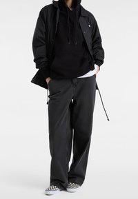 Giacca a vento nera sopra un hoodie nero con logo, abbinata a pantaloni neri larghi e scarpe slip-on a scacchi.