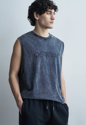 Νεαρός άνδρας με σγουρά μαλλιά, φορώντας μια ξεθωριασμένη γκρι αμάνικη μπλούζα "Off-White" και μαύρο παντελόνι, στέκεται με τα χέρια στις τσέπες, κοιτάζοντας προς τα δεξιά.