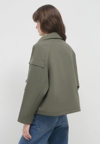 Femme portant une veste vert olive avec une poche à rabat sur la manches et un jean bleu, se tenant devant un fond blanc uni.