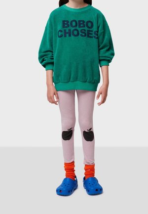 Niño vestido con un sudadera verde oversize con el texto "BOBO CHOSES", leggings rosa claro con estampados de manzanas negras en las rodillas, calcetines naranjas y zuecos azules.
