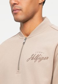Béžová mikina na zip s žebrovaným límcem a světle hnědým vyšitým názvem značky. Tkanina vypadá měkce s hladkým povrchem.