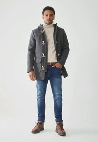 Uomo con cappotto grigio a toggle, maglione a collo alto beige in lavorazione a treccia, jeans blu e stivali marroni con lacci, in piedi contro uno sfondo semplice.