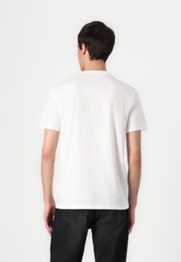Camiseta blanca de algodón con cuello redondo, mangas cortas y un corte clásico, mostrada desde la parte de atrás, contra un fondo liso.