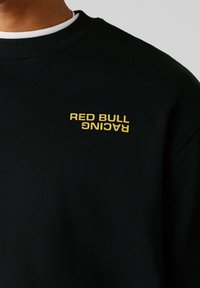 Osoba v čiernej mikine s žltým nápisom „RED BULL RACING“, kde je slovo „RACING“ na hrudi vytlačené obrátene.