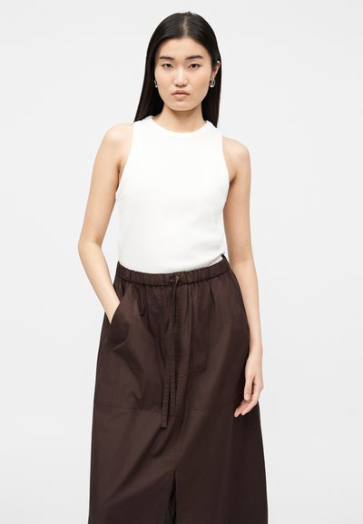 Jeune femme portant un haut blanc sans manches et une jupe taille haute marron foncé avec des poches, se tenant devant un fond clair uni.