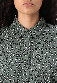 Gros plan d'une personne portant une chemise olive boutonnée avec un motif léopard noir et blanc.