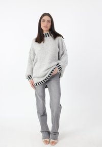 Grijze oversized gebreide trui met zwarte strepenaccenten bij de hals en de zoom, gecombineerd met lichtgrijze recht gesneden jeans en open teen sandalen.