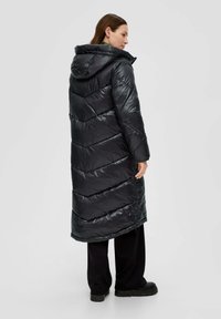Čierny dlhý puffer kabát s kapucňou, s designom chevron prešívania a lesklou textúrou, nosený cez čierne nohavice a robustné čierne čižmy.