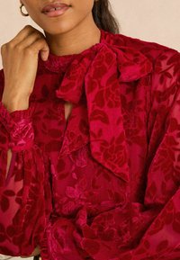 Röd blus med ett blommönster i sammet, hög hals, detaljer med rosett och samlade manschetter. Halvgennomsiktig tyg förstärker texturen.