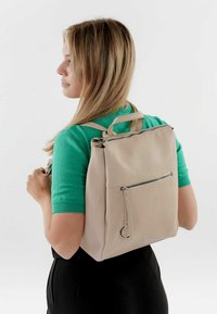 Beige ādas mugursoma ar augšējo rokturi, priekšējo rāvējslēdzēju kabatu un teksturētu apdari. Nēsāta uz muguras, apvienota ar zaļu augšu.