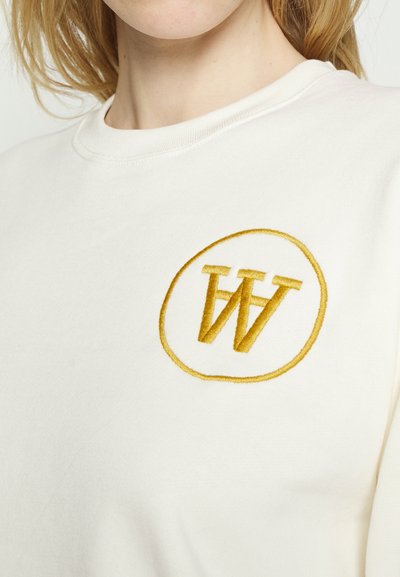 Hanorac crem cu un logo brodat circular galben, ce afișează literele interconectate "W" și "V". Textură moale și croială lejeră.