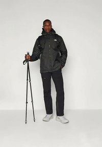 Must have veekindel must jope, must püksid ja valged saapad; hoiab matkakeppe; seljakott seljas.