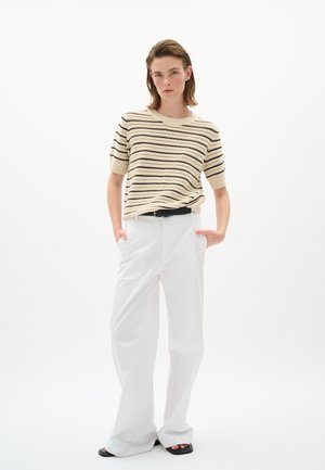 Donna che indossa un maglione a maniche corte beige e nero a righe, pantaloni bianchi a gamba larga con cintura nera e sandali neri, in piedi con le mani nelle tasche.