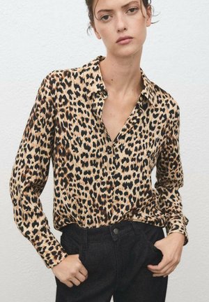 Woman wearing a long-sleeve leopard print blouse tucked into high-waist black pants, standing against a plain white wall.