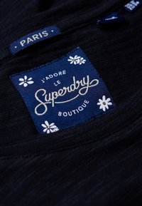 Svart tyg med en blå etikett med vita blommönster. Etiketten visar "Superdry", "J'adore le", och "Boutique, Paris."