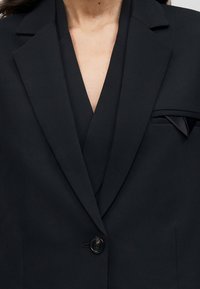 Μαύρο, καλοραμμένο σακάκι με κούμπωμα ενός κουμπιού, γιακά με χαραγμένη εγκοπή και λοξό μαντηλάκι στην τσέπη του στήθους.