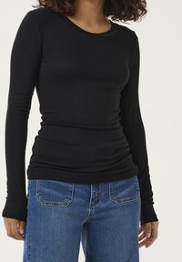 Woman wearing a fitted black long-sleeve shirt and blue denim jeans standing against a plain background.