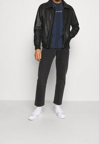 Svart läderbomberjacka, marinblå randig skjorta med logotyp, svarta slim-fit jeans, vita sneakers, stående mot en neutral bakgrund.