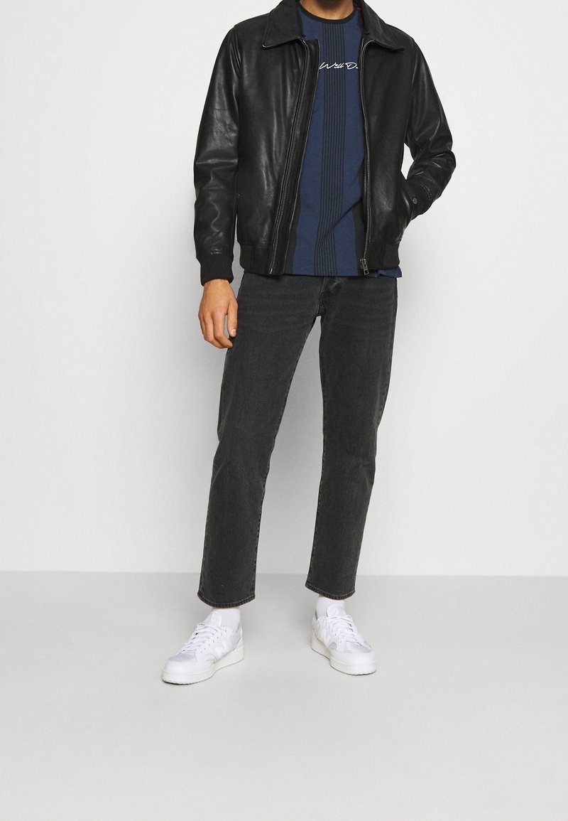 Svart läderbomberjacka, marinblå randig skjorta med logotyp, svarta slim-fit jeans, vita sneakers, stående mot en neutral bakgrund.