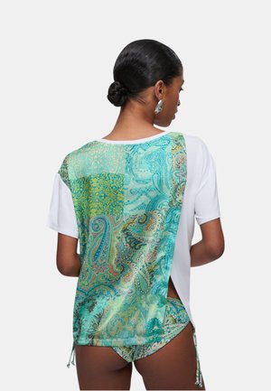 Donna con capelli scuri raccolti in uno chignon che indossa una maglietta bianca con un pannello posteriore paisley turchese colorato e slip bikini paisley abbinati.