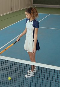Bílý a námořnický tenisový outfit se zkrácenou sukní a přiléhavým topem, stojící na modrém kurtu vedle tenisové sítě, držící raketu.