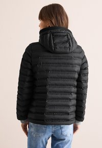 Veste doudoune noire avec capuche, présentant un design matelassé et une texture côtelée. Les manches affichent des poignets élastiques ; portée avec un jean bleu.