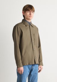 Mørk olivengrønn skjorte med krage, lange ermer og en enkelt brystlomme. Strukturert stoff kombinert med blå denimjeans.