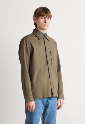Dark olive green button-up shirt with a collar, long sleeves, and a single chest pocket. Textured fabric paired with blue denim jeans.