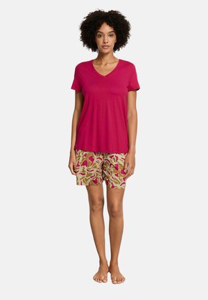 Kvinde stående barfodet iført en kortærmet rød V-hals trøje og beige shorts med røde og lyserøde abstrakte mønstre.