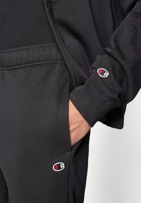 Μαύρο τζάκετ με φερμουάρ και παντελό νι από βαμβακερό μείγμα ύφασμα, με λογότυπο στο στήθος και στη μέση, λεία υφή χωρίς σχέδια.