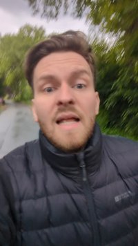 Giacca imbottita nera con zip e colletto rialzato. L'individuo ha capelli castano chiaro, una barba e si trova all'aperto con vegetazione sullo sfondo.