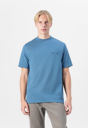 Blue short-sleeved t-shirt made of cotton with a crew neckline, featuring two small black logos on the chest, paired with light-colored pants.