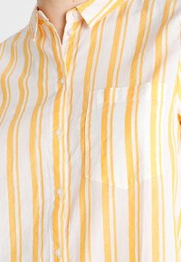 Camicia a maniche corte con strisce verticali gialle e bianche, caratterizzata da una chiusura con bottoni e una tasca sul petto. Tessuto morbido e leggero.