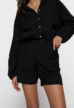 Kobieta w czarnej, zapinanej na guziki koszuli z długim rękawem, wpuszczonej w czarne, z wysokim stanem szorty z pofałdowaniami z przodu na jednolitym tle.