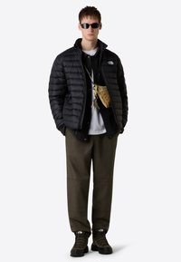 Giacca a vento nera, t-shirt bianca, camicia overshirt nera, pantaloni verde oliva, stivali neri e occhiali da sole. Gli accessori includono una piccola borsa beige.
