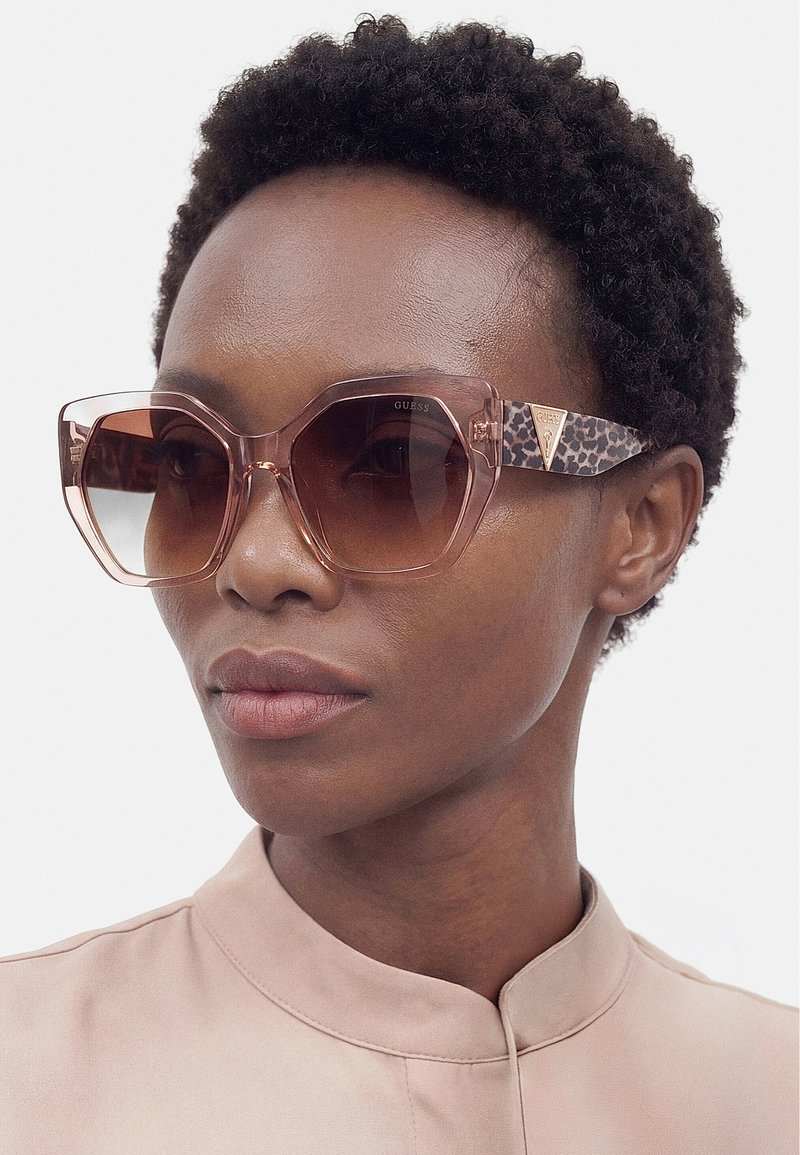 Femme aux cheveux courts et bouclés portant de grandes lunettes de soleil teintées roses avec des branches imprimées léopard et un haut beige sans col.