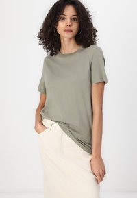Olive green cotton t-shirt with short sleeves, relaxed fit, and a round neckline, paired with a light beige skirt.