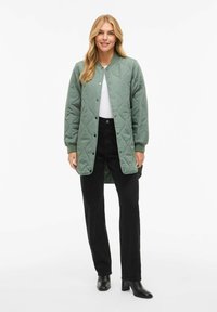 Su quiltingas, vidutinio ilgio žalias paltas su apvalia apykakle ir spaustukais, derinamas su juodomis kelnėmis ir juodais aukštakulniais batais.