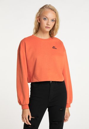 Junge Frau mit blonden Haaren trägt ein oranges Crop-Sweatshirt mit dem Schriftzug „REBEL with a cause“ und schwarze, zerrissene Skinny-Jeans.