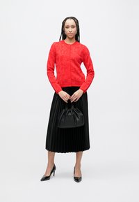 Rode gebreide cardigan met bloemenpatroon, gecombineerd met een plissé zwarte rok. Model draagt een zwarte leren trekkoordtas en zwarte hakken.