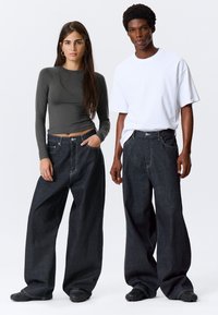 Model nosi prilagođenu sivo dugom rukavu majicu, uparen sa prevelikim tamnim trapericama. Traperice imaju klasičan dizajn s pet džepova i vidljivim šavovima.